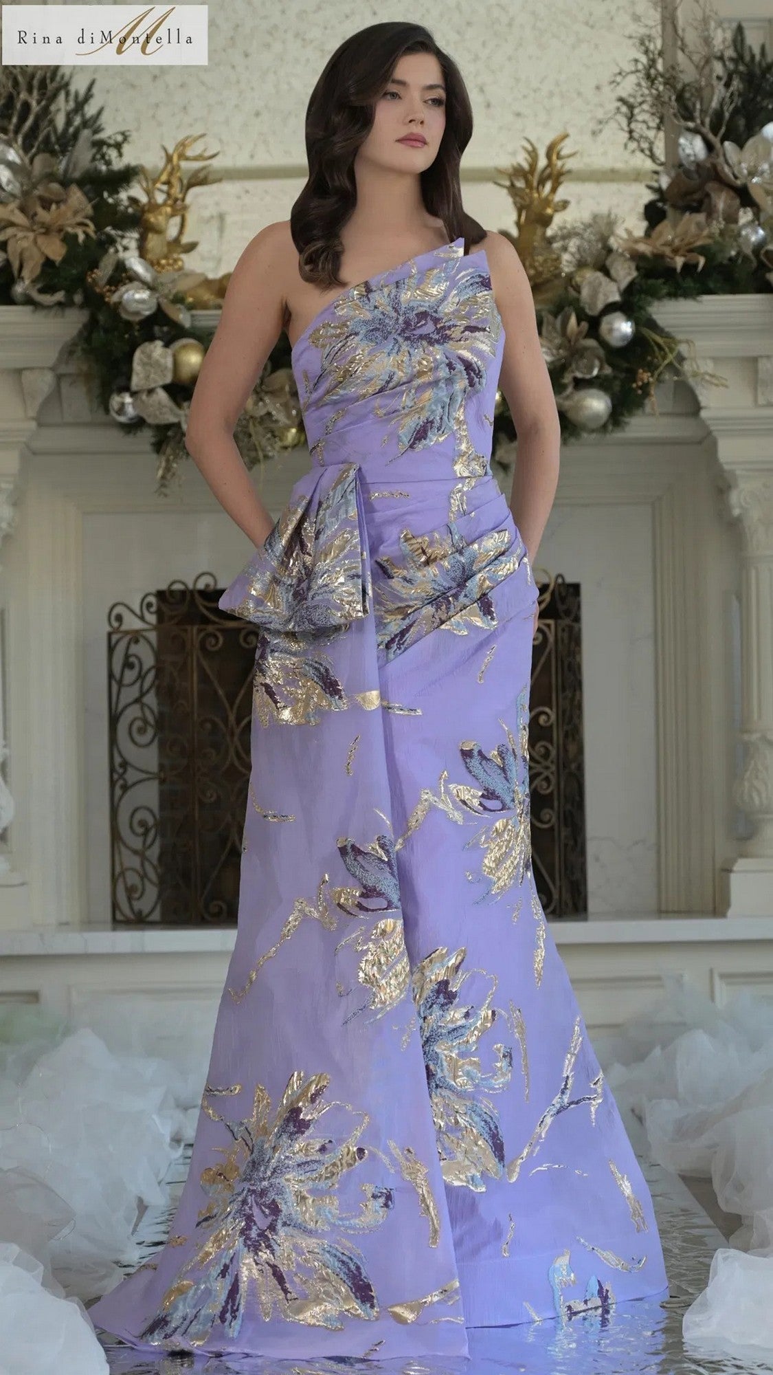 A woman stands indoors by a decorated fireplace wearing the Rina Di Montella RD3233, a long formal one-shoulder dress in Lavender Mu with gold and silver floral organza jacquard patterns.